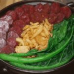 Top-down view of cooked claypot rice with sliced Chinese sausage, vibrant green choy sum, and crispy shallots arranged neatly in a clay pot, highlighting the textures and colors of the final dish.