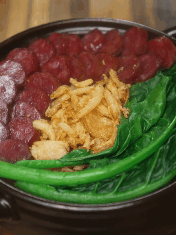 Top-down view of cooked claypot rice with sliced Chinese sausage, vibrant green choy sum, and crispy shallots arranged neatly in a clay pot, highlighting the textures and colors of the final dish.