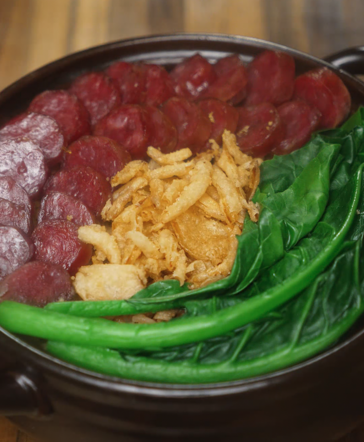Top-down view of cooked claypot rice with sliced Chinese sausage, vibrant green choy sum, and crispy shallots arranged neatly in a clay pot, highlighting the textures and colors of the final dish.