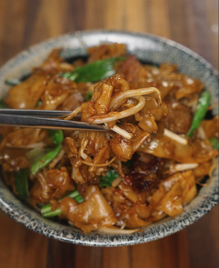 A close-up of glossy Beef Chow Fun with chopsticks lifting a perfect bite of noodles, beef, and bean sprouts. This image highlights the savory glaze and ideal texture balance.