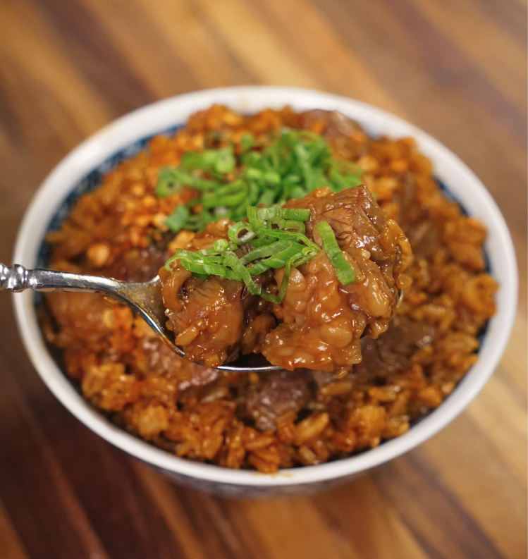 A spoonful of garlic steak fried rice being lifted from a full bowl, showcasing the glossy beef, crispy garlic, and green onion garnish in a close-up view.