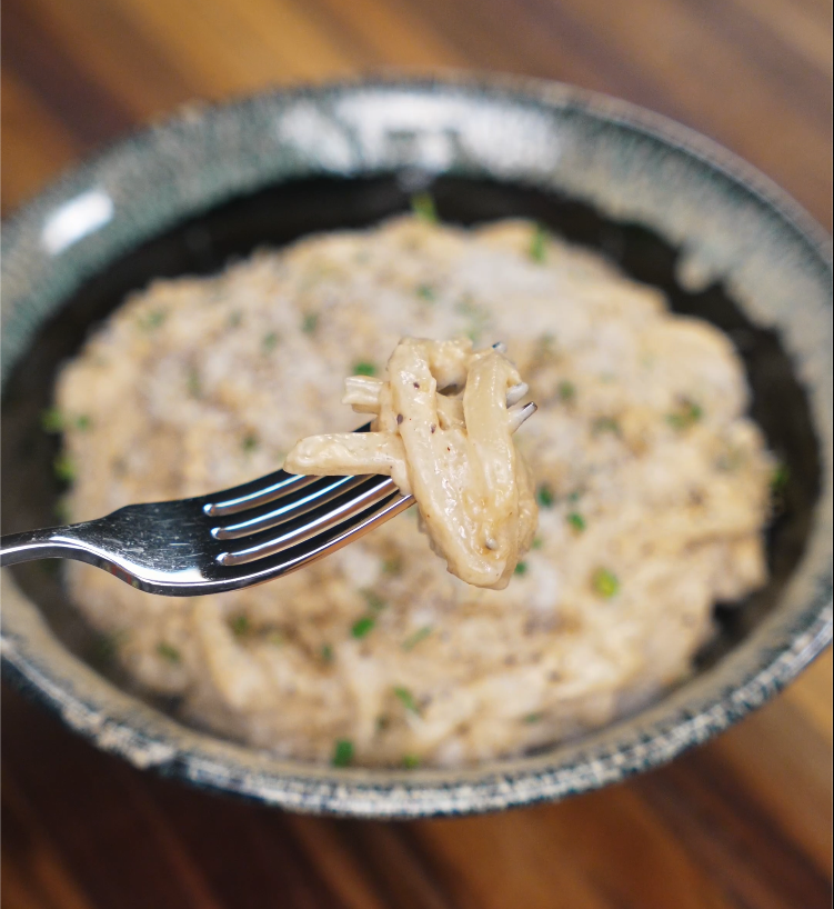 A fork lifting a bite of creamy garlic noodles from the bowl, showcasing the sauce's silky texture and garnishes in the background.