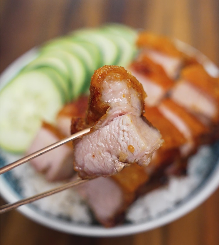 A single piece of crispy pork belly held by chopsticks above a bowl of rice, showcasing its juicy interior and golden crackling skin.