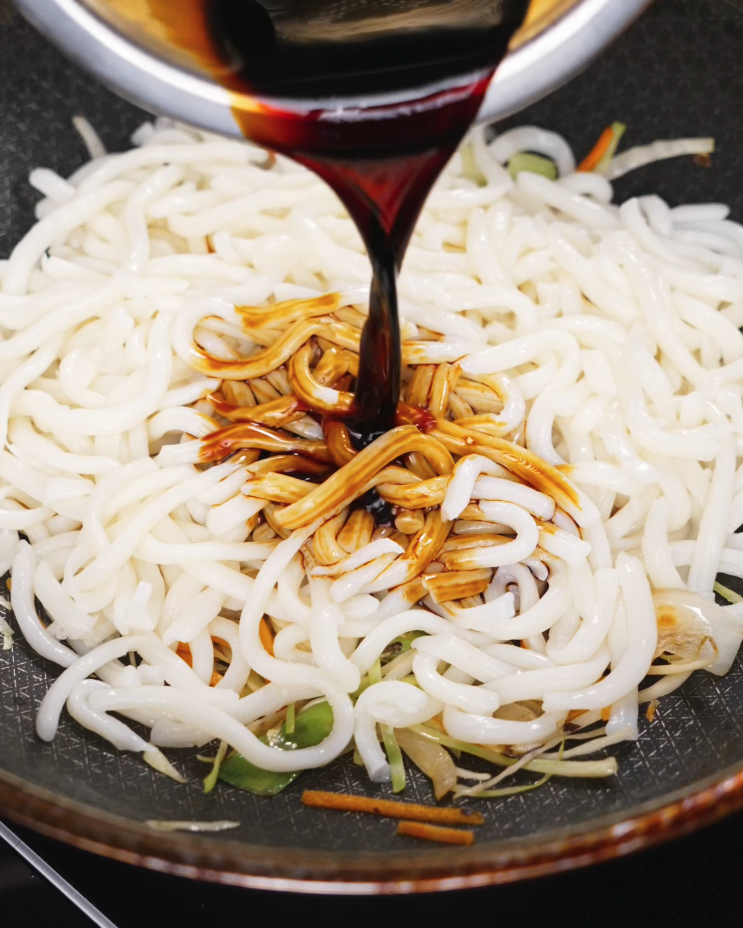 Dark soy-based sauce being poured over stir-fried udon noodles and vegetables in a non-stick skillet, showing the beginning of the dish’s signature glossy glaze.