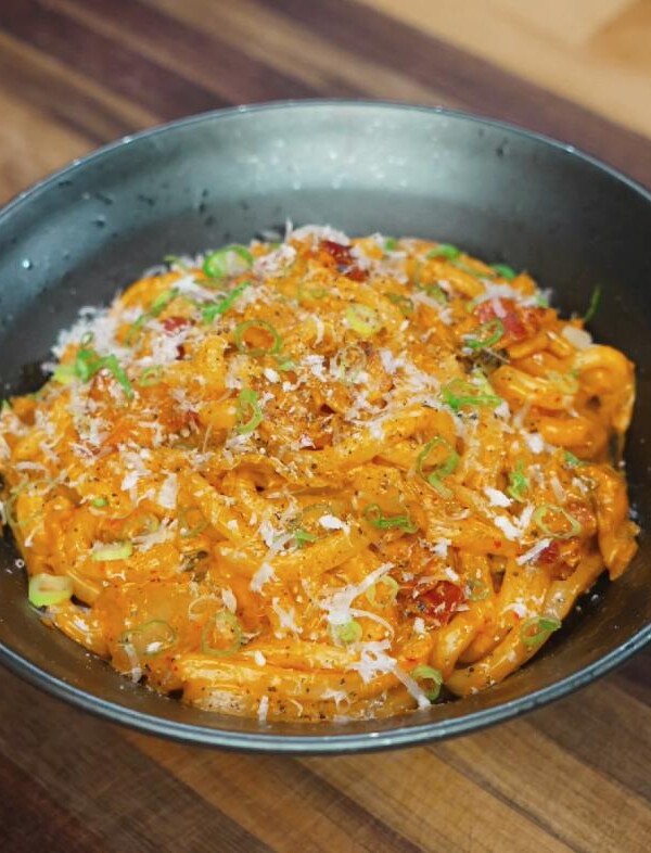 Close-up of creamy kimchi noodles served in a black bowl, topped with scallions, chili flakes, and shredded cheese for garnish.