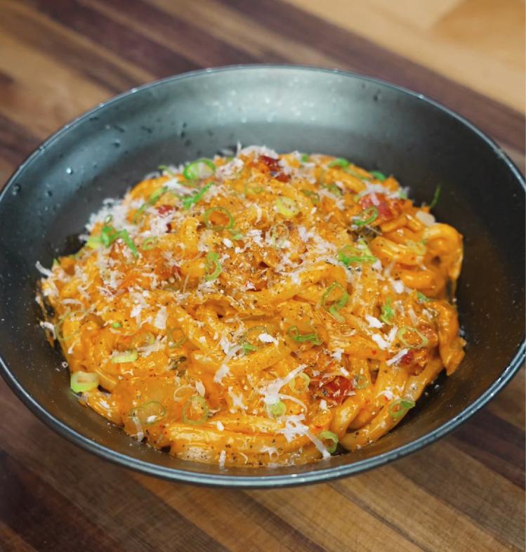 Close-up of creamy kimchi noodles served in a black bowl, topped with scallions, chili flakes, and shredded cheese for garnish.