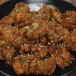 Crispy honey garlic chicken served in a black bowl, topped with sesame seeds and thinly sliced green onions. The chicken is golden brown and coated in a sticky glaze.