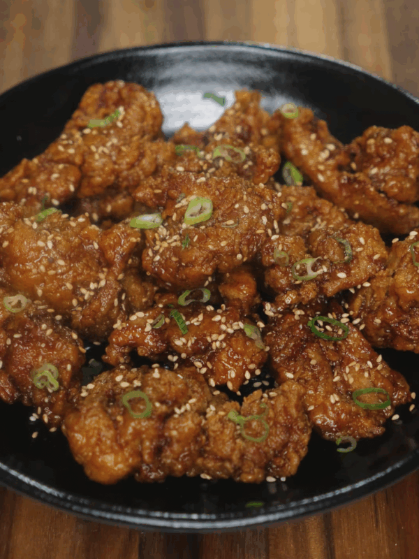 Crispy honey garlic chicken served in a black bowl, topped with sesame seeds and thinly sliced green onions. The chicken is golden brown and coated in a sticky glaze.