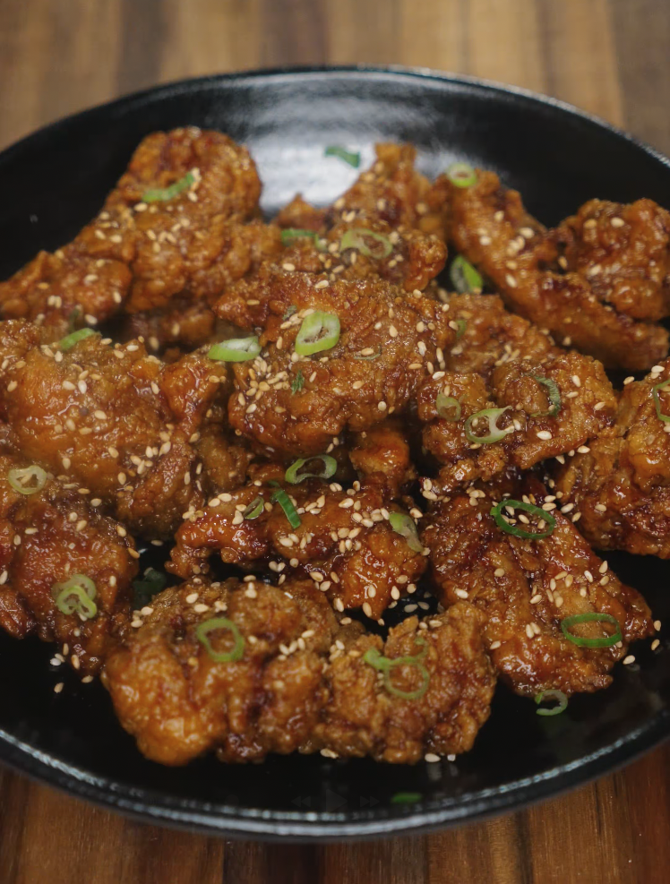 Crispy honey garlic chicken served in a black bowl, topped with sesame seeds and thinly sliced green onions. The chicken is golden brown and coated in a sticky glaze.
