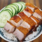 Crispy pork belly sliced into thick pieces, served over a bed of steamed jasmine rice with cucumber slices, all placed in a blue and white patterned bowl.