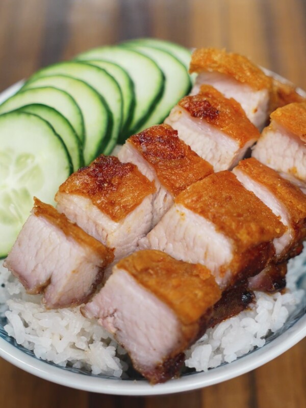 Crispy pork belly sliced into thick pieces, served over a bed of steamed jasmine rice with cucumber slices, all placed in a blue and white patterned bowl.