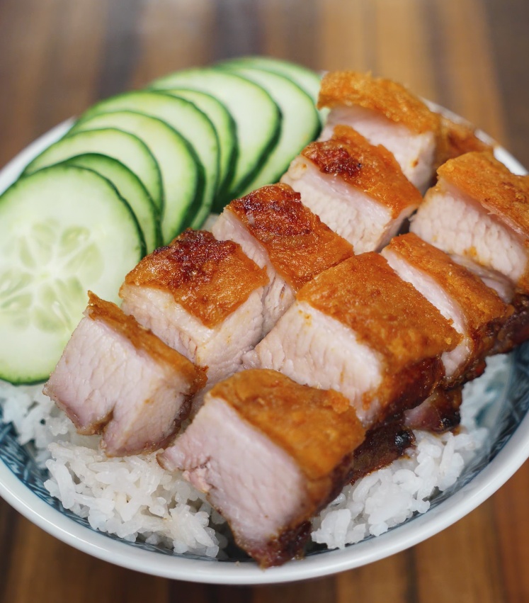 Crispy pork belly sliced into thick pieces, served over a bed of steamed jasmine rice with cucumber slices, all placed in a blue and white patterned bowl.