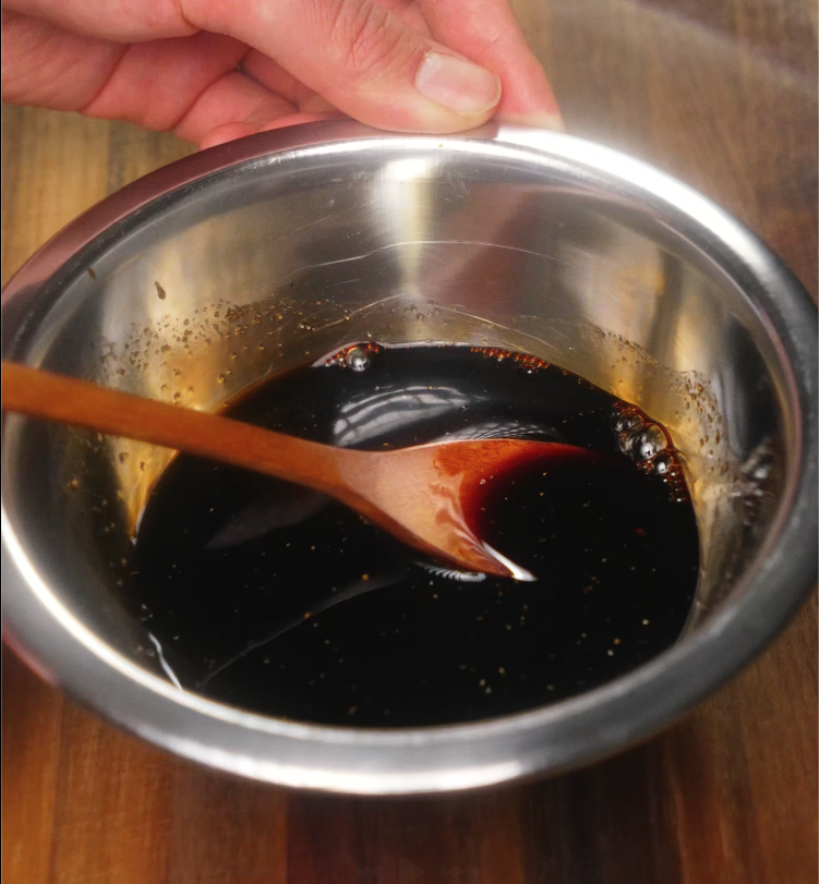 Close-up of a dark soy sauce mixture being stirred in a stainless steel bowl with a wooden spoon, showing the glossy and rich sauce that will flavor the claypot rice.