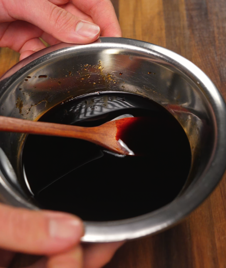 A rich, glossy soy-based sauce being stirred in a metal mixing bowl with a wooden spoon, preparing the flavorful stir-fry base.