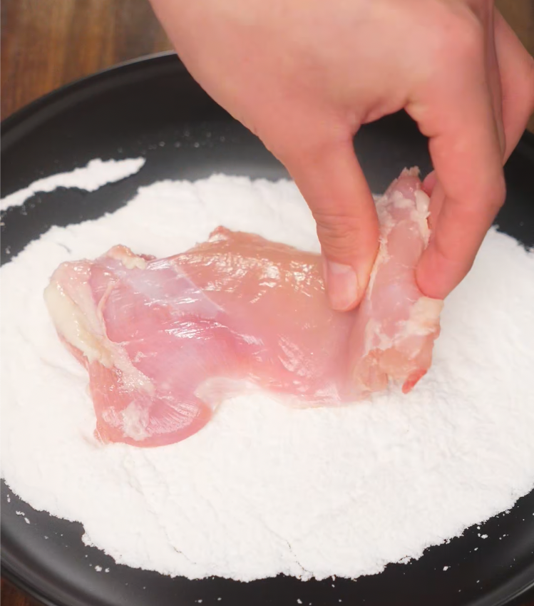 Raw boneless chicken thigh being dredged in flour as the first step in preparing crispy katsu.