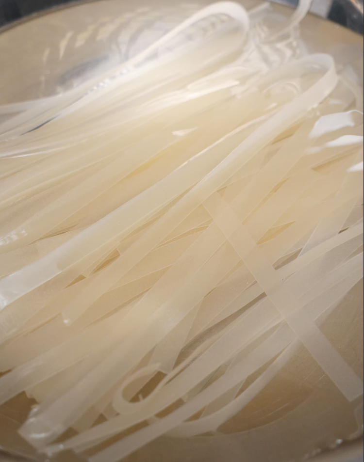 Close-up of dry, uncooked wide rice noodles sitting in a glass bowl, showing their translucent, stiff texture before soaking or boiling.
