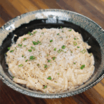 A bowl of creamy garlic noodles topped with grated parmesan, black pepper, and chopped scallions, served in a rustic green-edged ceramic bowl.