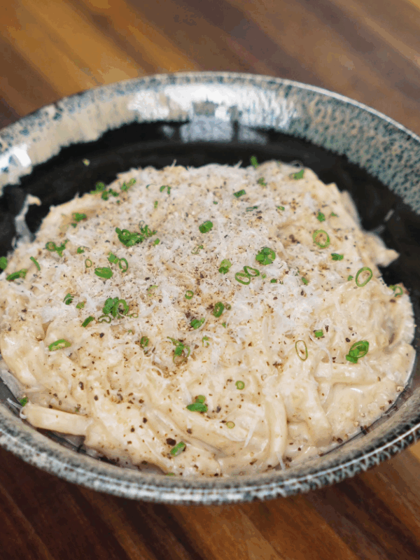 A bowl of creamy garlic noodles topped with grated parmesan, black pepper, and chopped scallions, served in a rustic green-edged ceramic bowl.