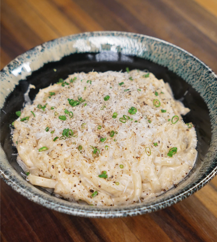 A bowl of creamy garlic noodles topped with grated parmesan, black pepper, and chopped scallions, served in a rustic green-edged ceramic bowl.