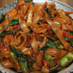 A bowl of drunken noodles piled high with stir-fried wide rice noodles, vibrant green Chinese broccoli, red chili, carrots, and tender chicken pieces, all coated in a glossy savory sauce and garnished with Thai basil.