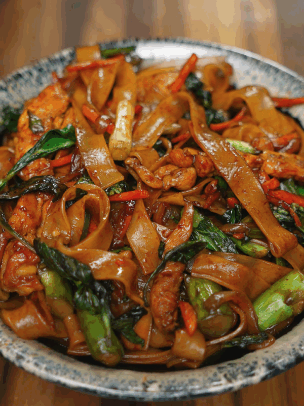 A bowl of drunken noodles piled high with stir-fried wide rice noodles, vibrant green Chinese broccoli, red chili, carrots, and tender chicken pieces, all coated in a glossy savory sauce and garnished with Thai basil.