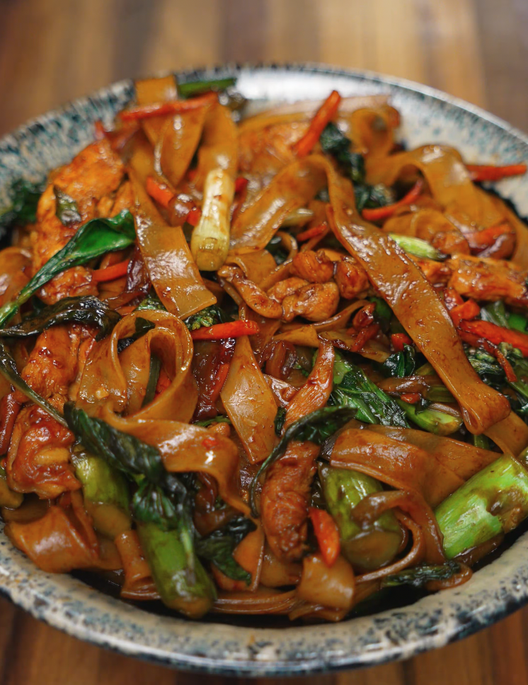 A bowl of drunken noodles piled high with stir-fried wide rice noodles, vibrant green Chinese broccoli, red chili, carrots, and tender chicken pieces, all coated in a glossy savory sauce and garnished with Thai basil.