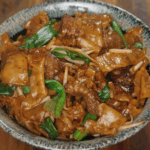 A finished bowl of Beef Chow Fun served in a speckled ceramic dish, featuring glossy stir-fried rice noodles, tender beef, scallions, and bean sprouts coated in a rich soy-based sauce.