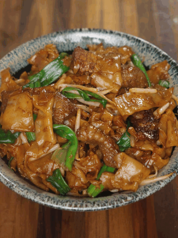 A finished bowl of Beef Chow Fun served in a speckled ceramic dish, featuring glossy stir-fried rice noodles, tender beef, scallions, and bean sprouts coated in a rich soy-based sauce.