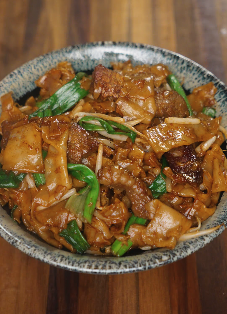 A finished bowl of Beef Chow Fun served in a speckled ceramic dish, featuring glossy stir-fried rice noodles, tender beef, scallions, and bean sprouts coated in a rich soy-based sauce.