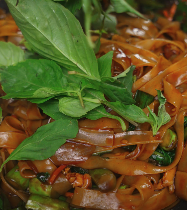 A generous handful of fresh Thai basil leaves being scattered on top of stir-fried drunken noodles, ready to wilt into the hot dish.