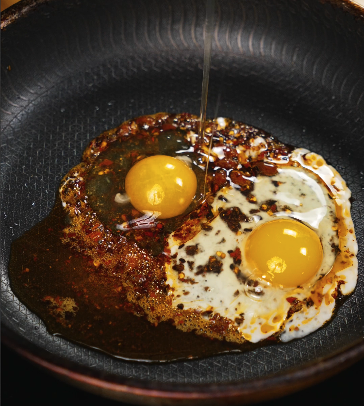Two sunny side up eggs frying in chili oil in a nonstick skillet, yolks intact and surrounded by crisped chili flakes.