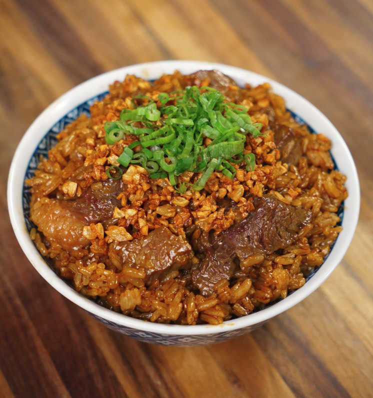 A high-resolution photo of garlic steak fried rice served in a blue and white patterned bowl, garnished with chopped scallions on top of crispy garlic and glossy beef pieces for a flavorful finish.