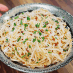 A bowl of garlic butter noodles topped with shredded parmesan cheese, red chili flakes, and finely chopped parsley, served on a wooden surface.