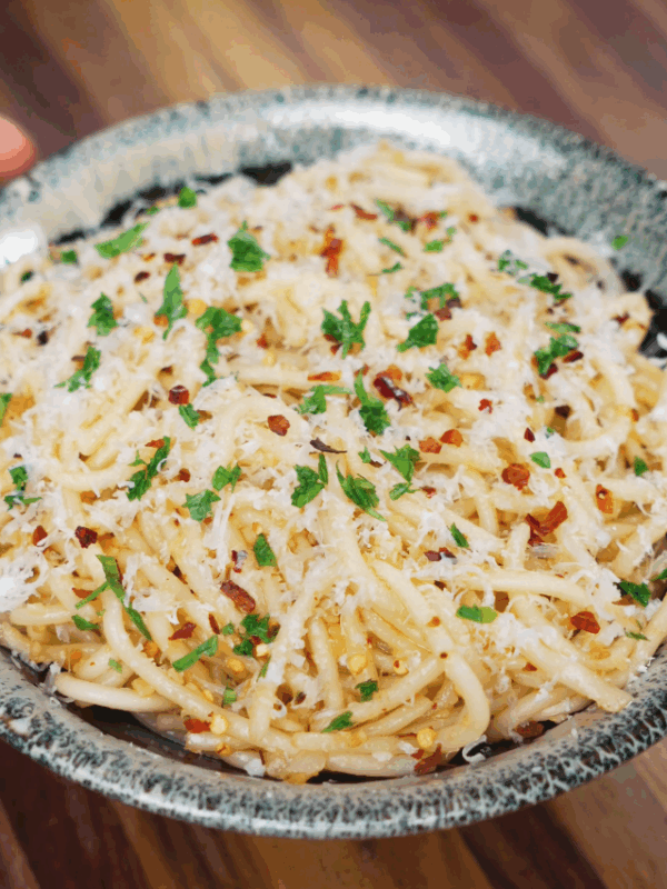 A bowl of garlic butter noodles topped with shredded parmesan cheese, red chili flakes, and finely chopped parsley, served on a wooden surface.
