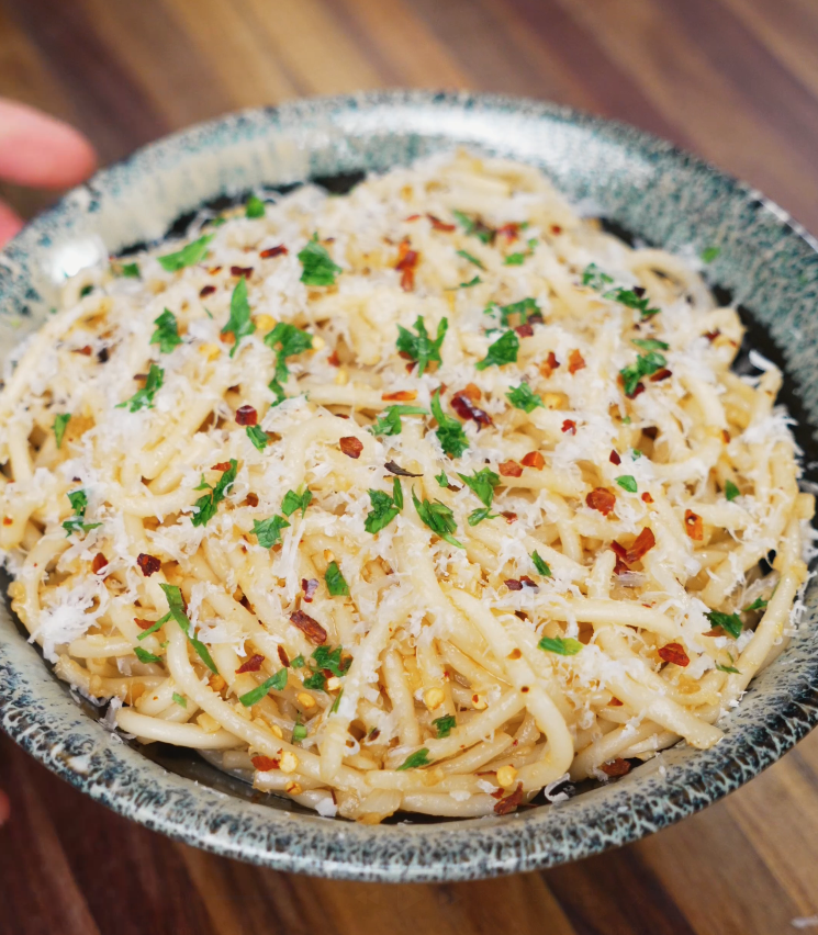 A bowl of garlic butter noodles topped with shredded parmesan cheese, red chili flakes, and finely chopped parsley, served on a wooden surface.