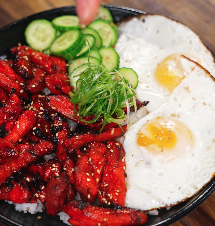 Garnishing the finished Chicken Tocino bowl with thinly sliced scallions and crisp cucumbers. The vibrant toppings add freshness and crunch to the rich, savory meal.