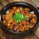 A high-resolution image shows ginger braised chicken served over white rice in a black ceramic bowl, garnished with chopped scallions. The glossy glaze of the chicken contrasts beautifully with the wooden surface beneath the bowl.