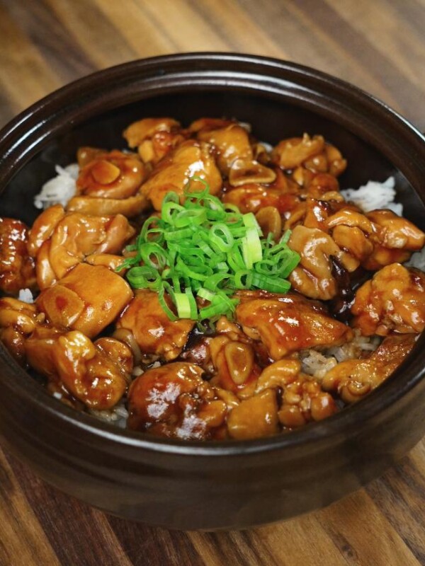 A high-resolution image shows ginger braised chicken served over white rice in a black ceramic bowl, garnished with chopped scallions. The glossy glaze of the chicken contrasts beautifully with the wooden surface beneath the bowl.