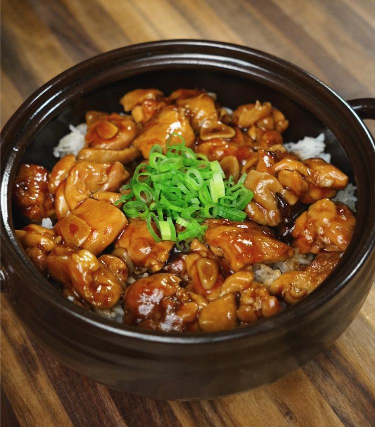 A high-resolution image shows ginger braised chicken served over white rice in a black ceramic bowl, garnished with chopped scallions. The glossy glaze of the chicken contrasts beautifully with the wooden surface beneath the bowl.