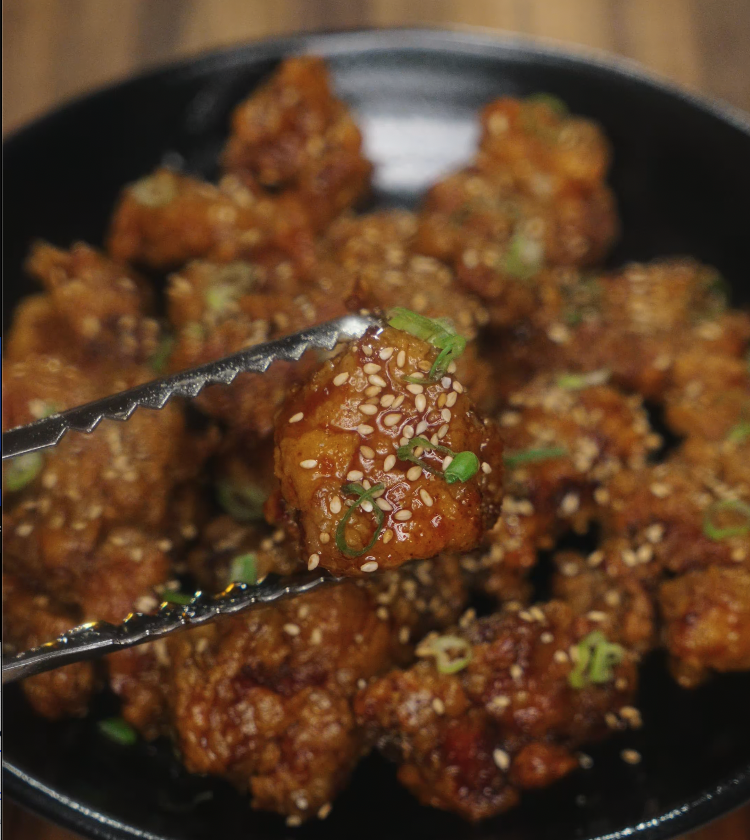 Tongs lifting a piece of honey garlic chicken showing the sticky glaze, sesame seeds, and scallion garnish.