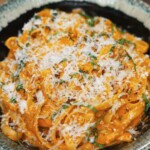 A close-up shot of Gochujang Alfredo noodles served in a speckled ceramic bowl, generously topped with freshly grated parmesan and chopped herbs, highlighting the creamy, spicy sauce coating each strand.