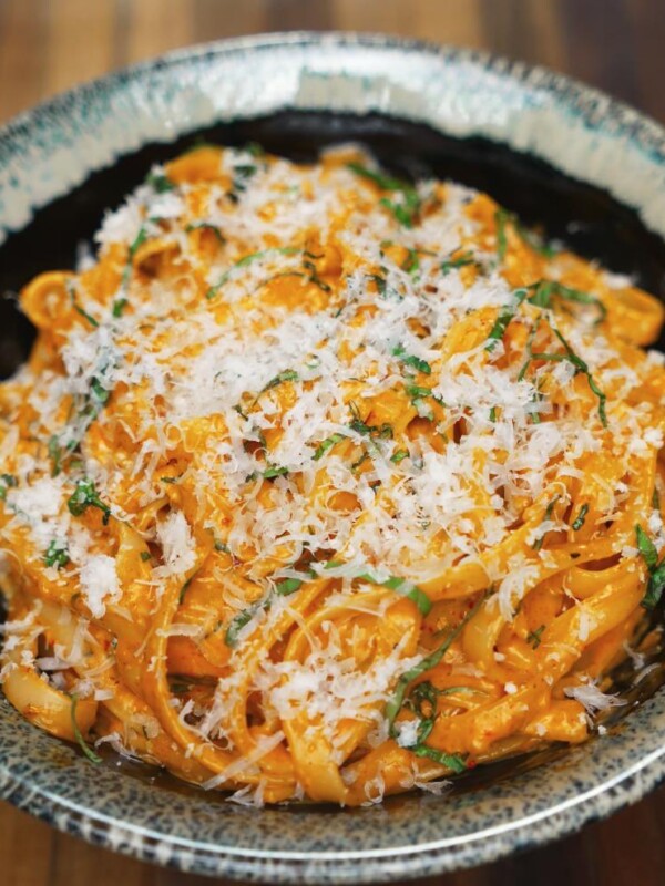 A close-up shot of Gochujang Alfredo noodles served in a speckled ceramic bowl, generously topped with freshly grated parmesan and chopped herbs, highlighting the creamy, spicy sauce coating each strand.
