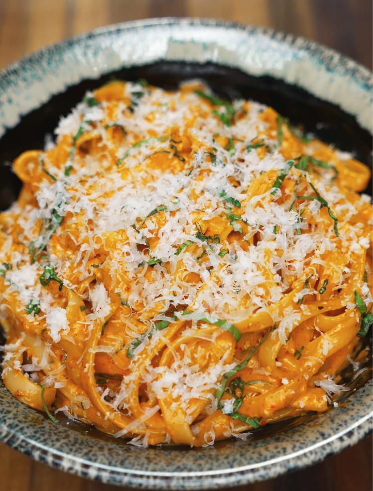 A close-up shot of Gochujang Alfredo noodles served in a speckled ceramic bowl, generously topped with freshly grated parmesan and chopped herbs, highlighting the creamy, spicy sauce coating each strand.