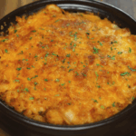 A golden-brown gochujang mac and cheese baked in a brown ceramic dish, topped with broiled melted cheese and sprinkled with finely chopped herbs for garnish.