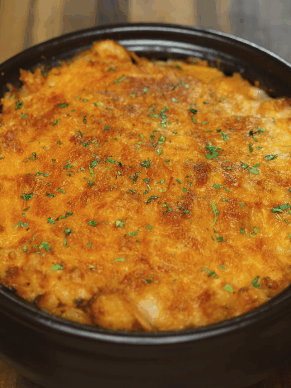 A golden-brown gochujang mac and cheese baked in a brown ceramic dish, topped with broiled melted cheese and sprinkled with finely chopped herbs for garnish.