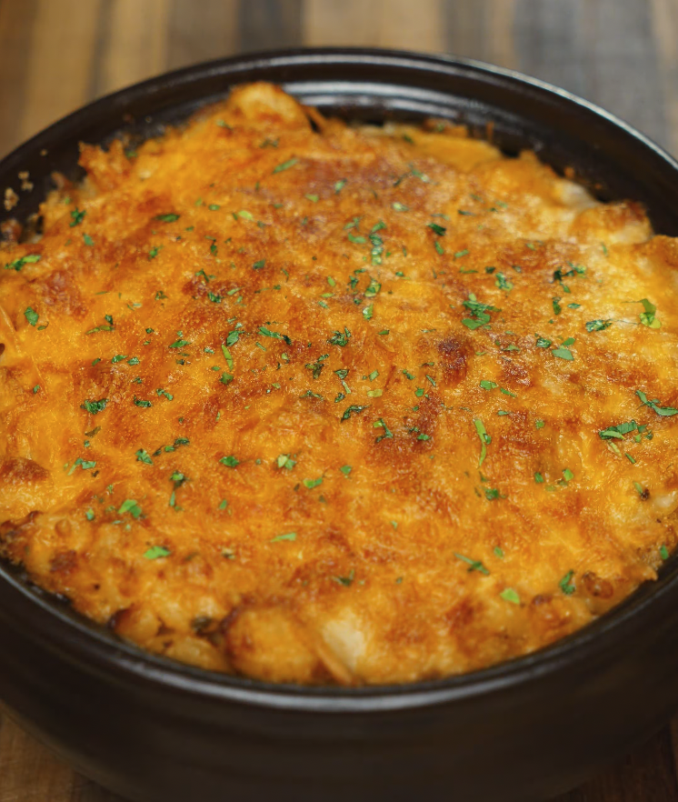 A golden-brown gochujang mac and cheese baked in a brown ceramic dish, topped with broiled melted cheese and sprinkled with finely chopped herbs for garnish.