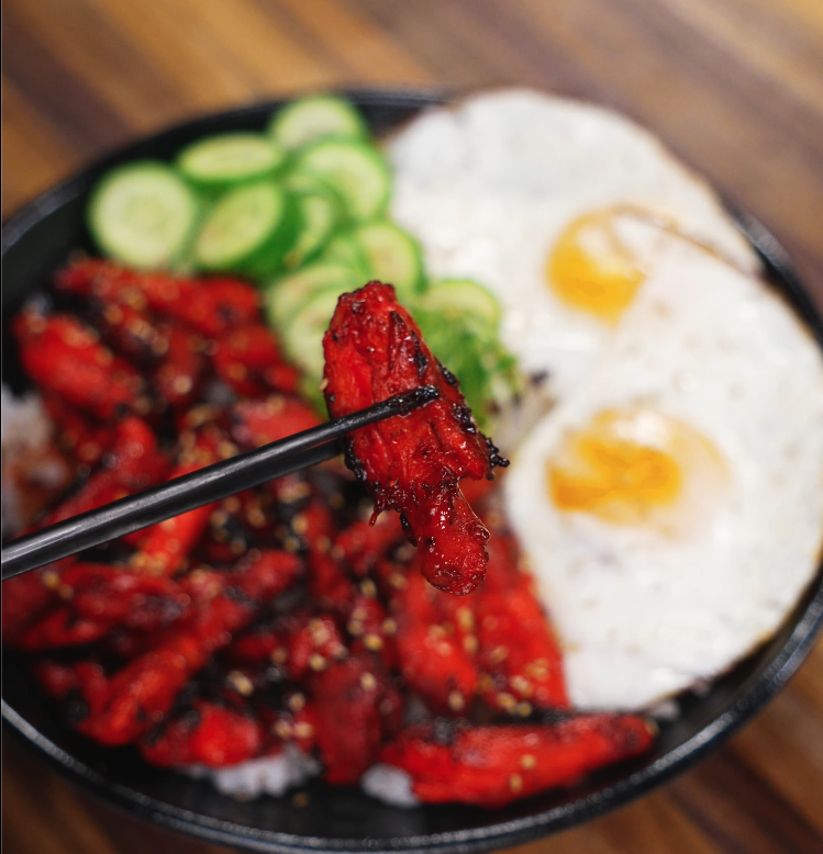 Close-up of a piece of glazed Chicken Tocino held with chopsticks over a bowl of rice, eggs, and vegetables. The sticky, sweet-savory exterior highlights the caramelization that makes tocino irresistible.
