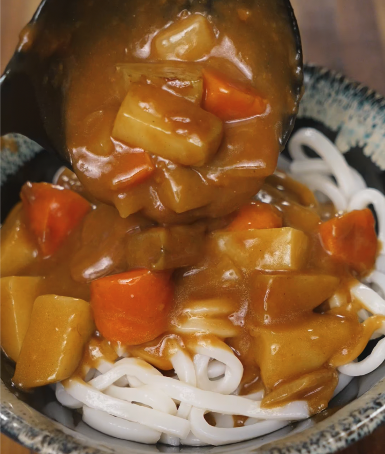 Hearty Japanese curry with vegetables being ladled generously over a bowl of udon noodles.