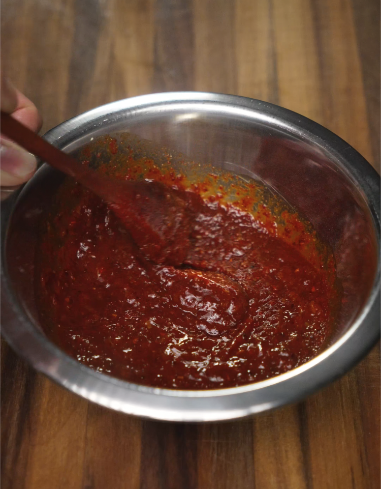 Stirring together gochujang, soy sauce, garlic, and chili flakes in a metal bowl to make the spicy soup base for Korean army stew.