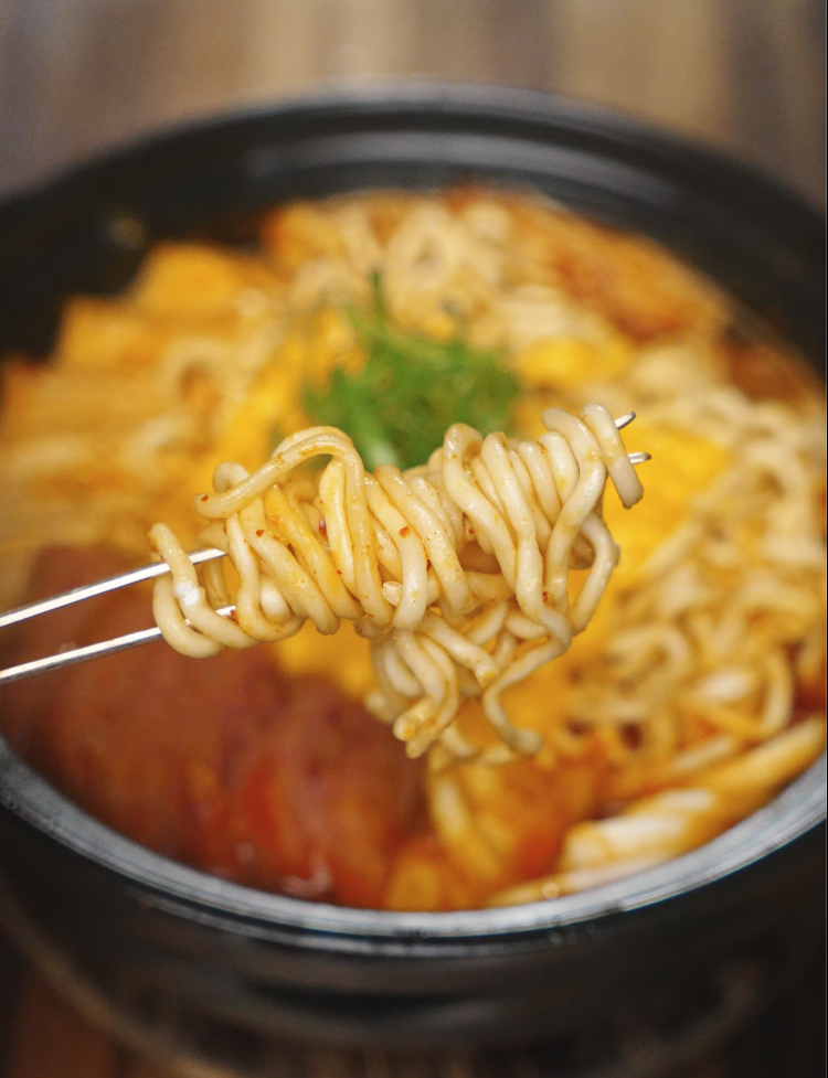 Close-up shot of metal chopsticks lifting perfectly cooked ramen noodles from the center of the bubbling budae jjigae.